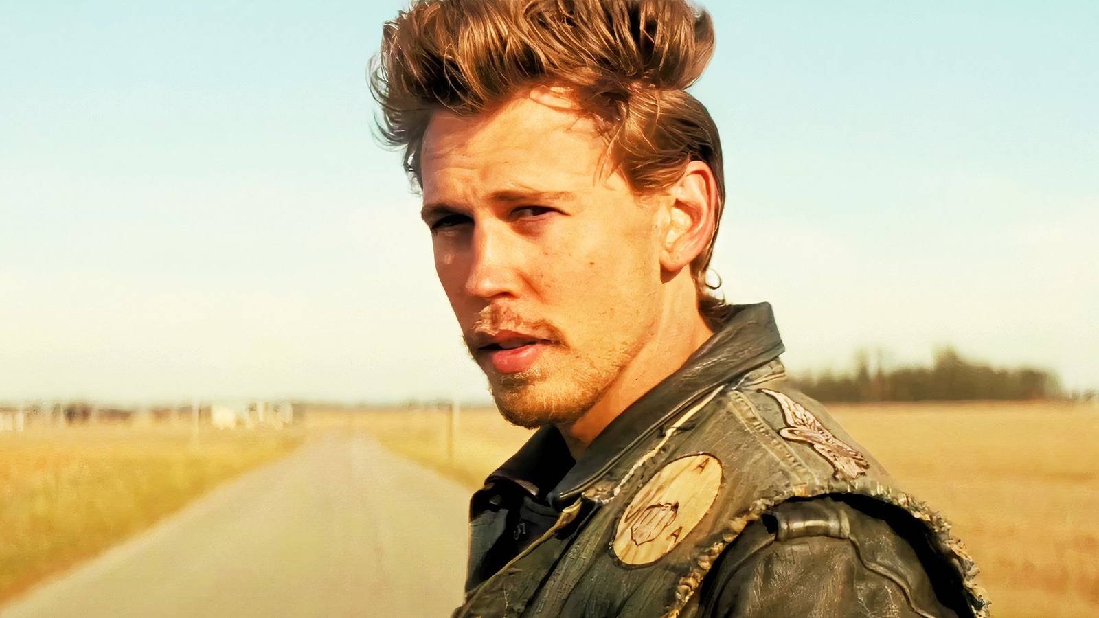 Austin Butler as Benny in The Bikeriders standing on a country road.