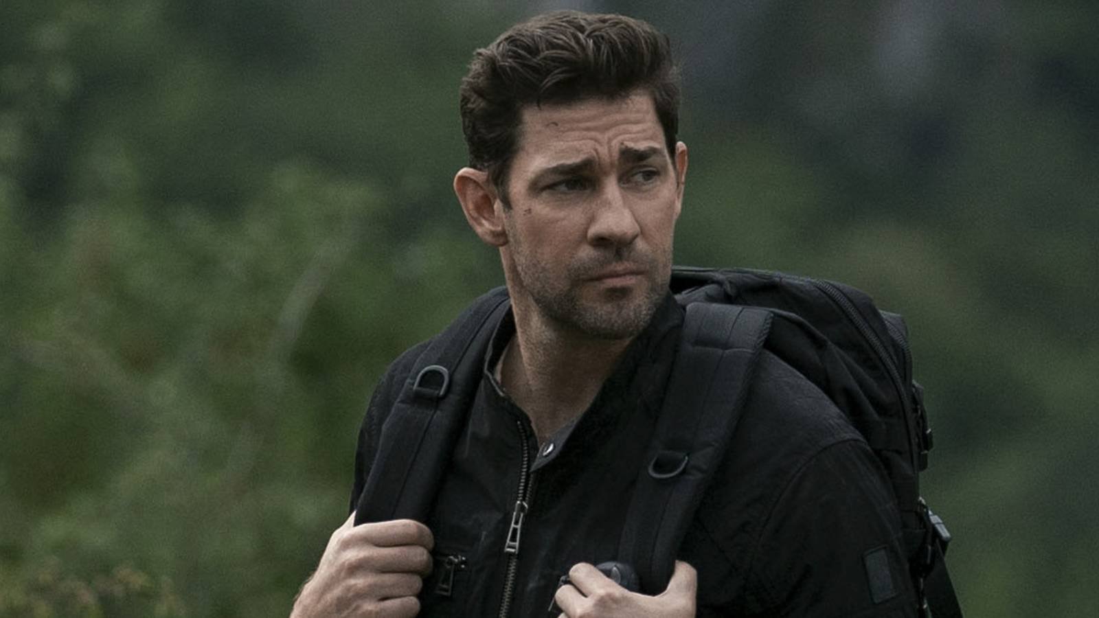 John Krasinski as Jack Ryan walking in front of some greenery while holding the straps of his bac...